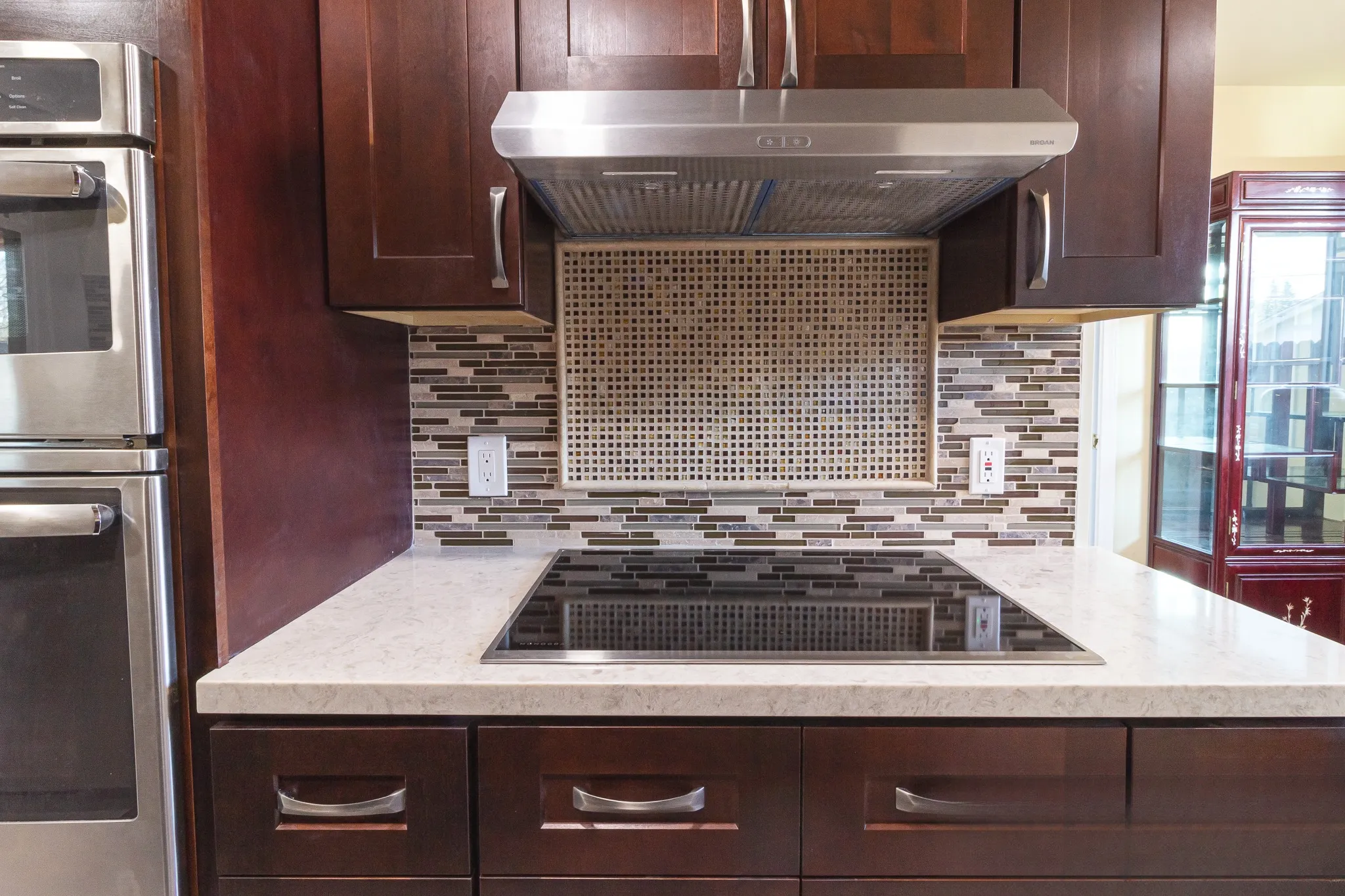 Cooktop below a stainless hood with a square mosaic inset panel set into the linear glass mosaic backsplash