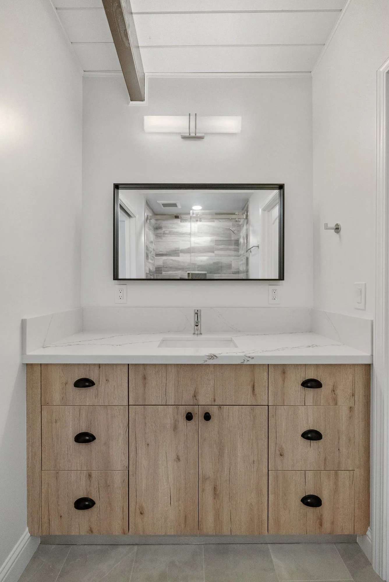 Same bathroom after remodel, light oak double vanity with quartz top, black hardware and a black-framed mirror
