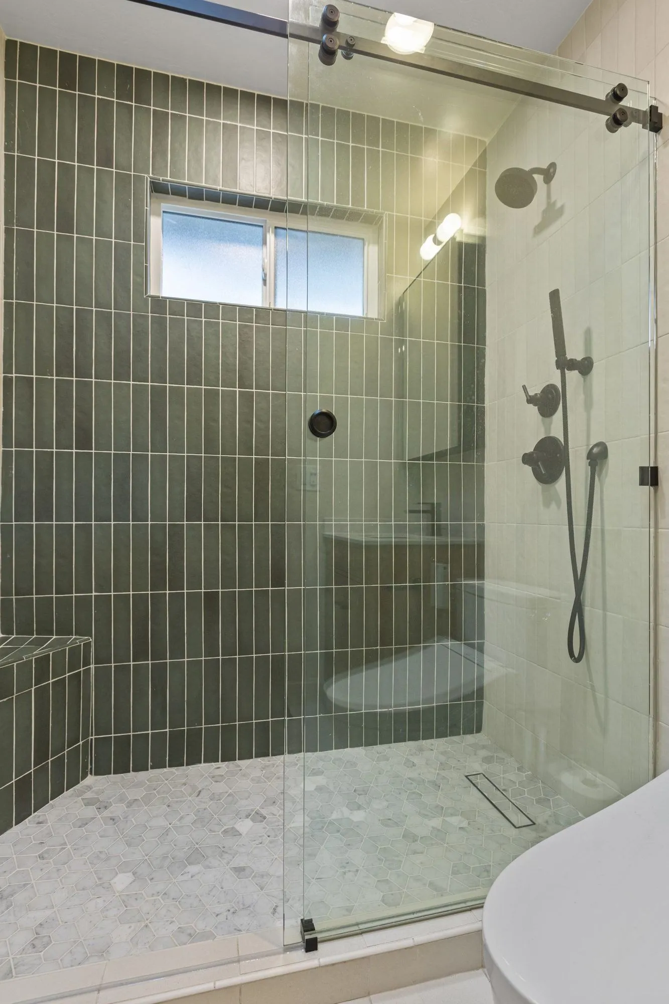 Shower behind a frameless glass screen, sage-green stacked tile with a pebble mosaic floor