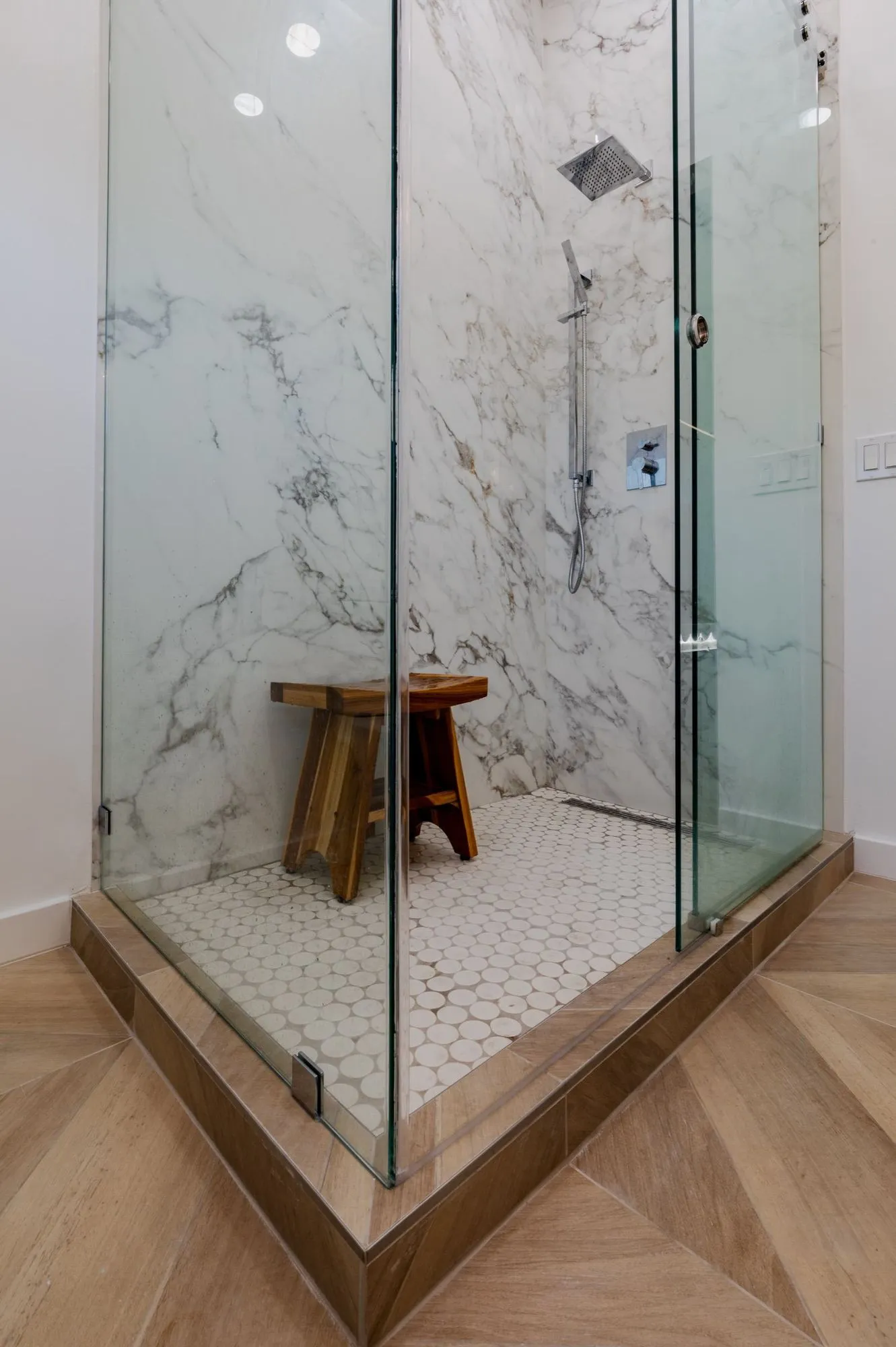 Frameless glass shower after remodel, marble-veined slab walls with white hex tile floor and teak stool