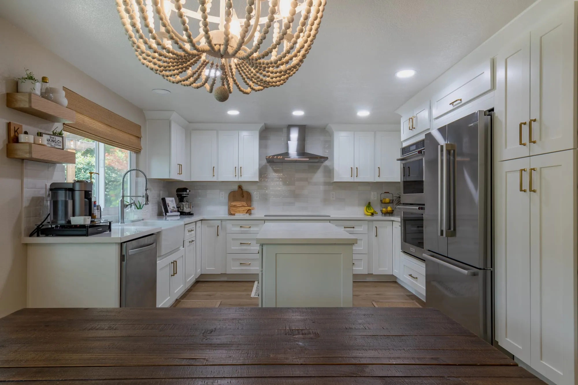 Kitchen after remodel, white shaker cabinetry with brass pulls around a sage-green island under a beaded chandelier