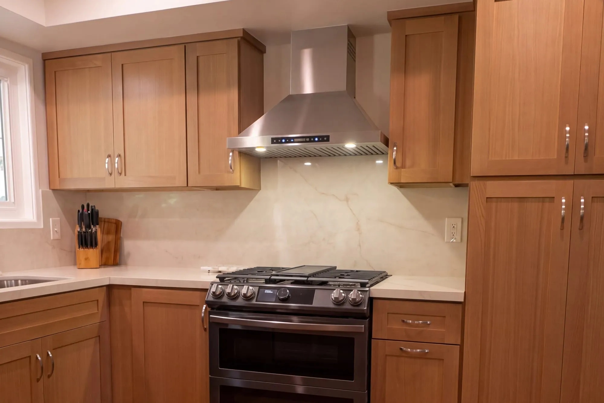 Kitchen after remodel, wood shaker cabinetry under a full-height quartz backsplash with a stainless chimney hood above the range
