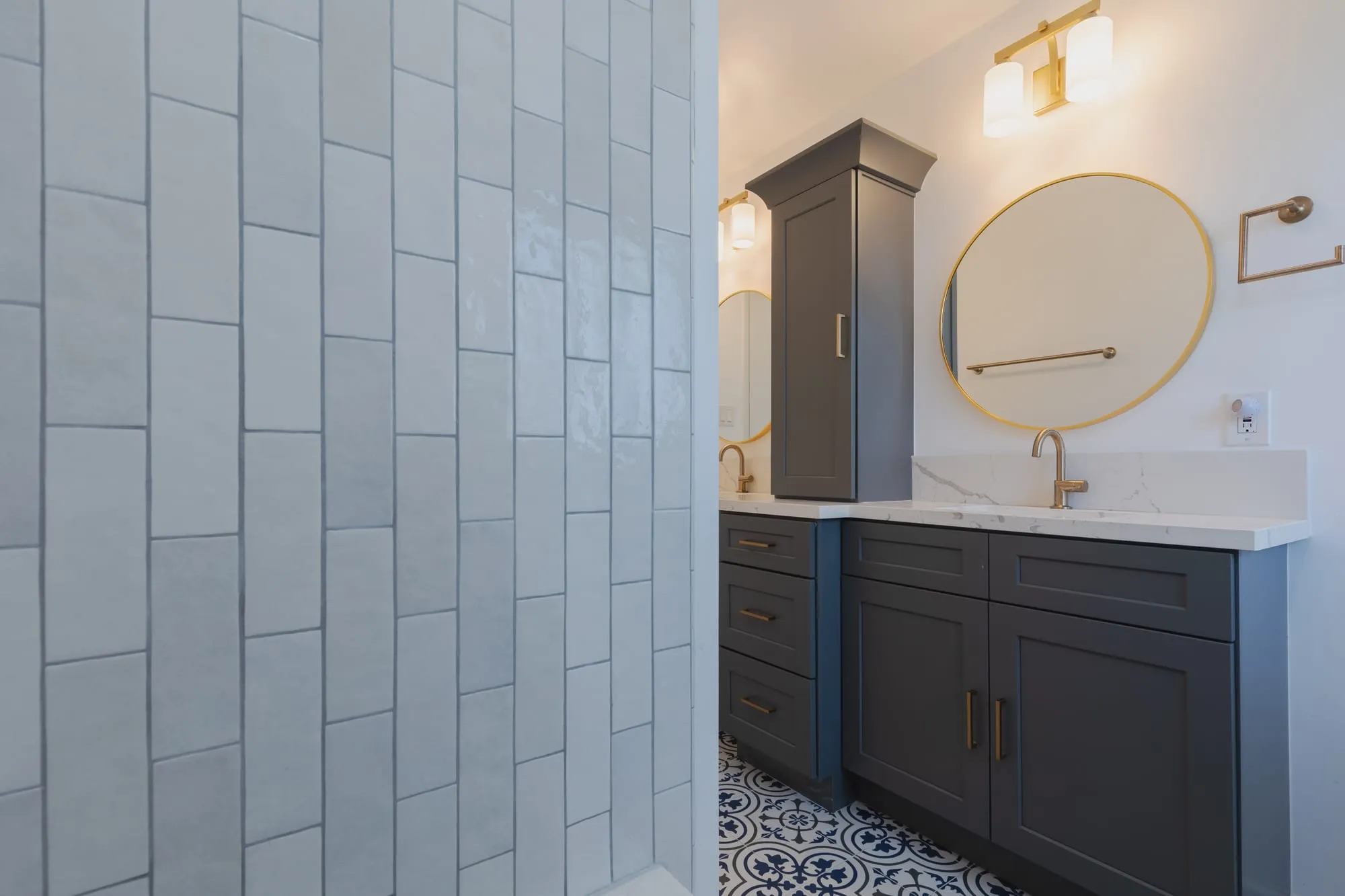 Bathroom after remodel, grey shaker vanity under a round brass mirror beside stacked white tile