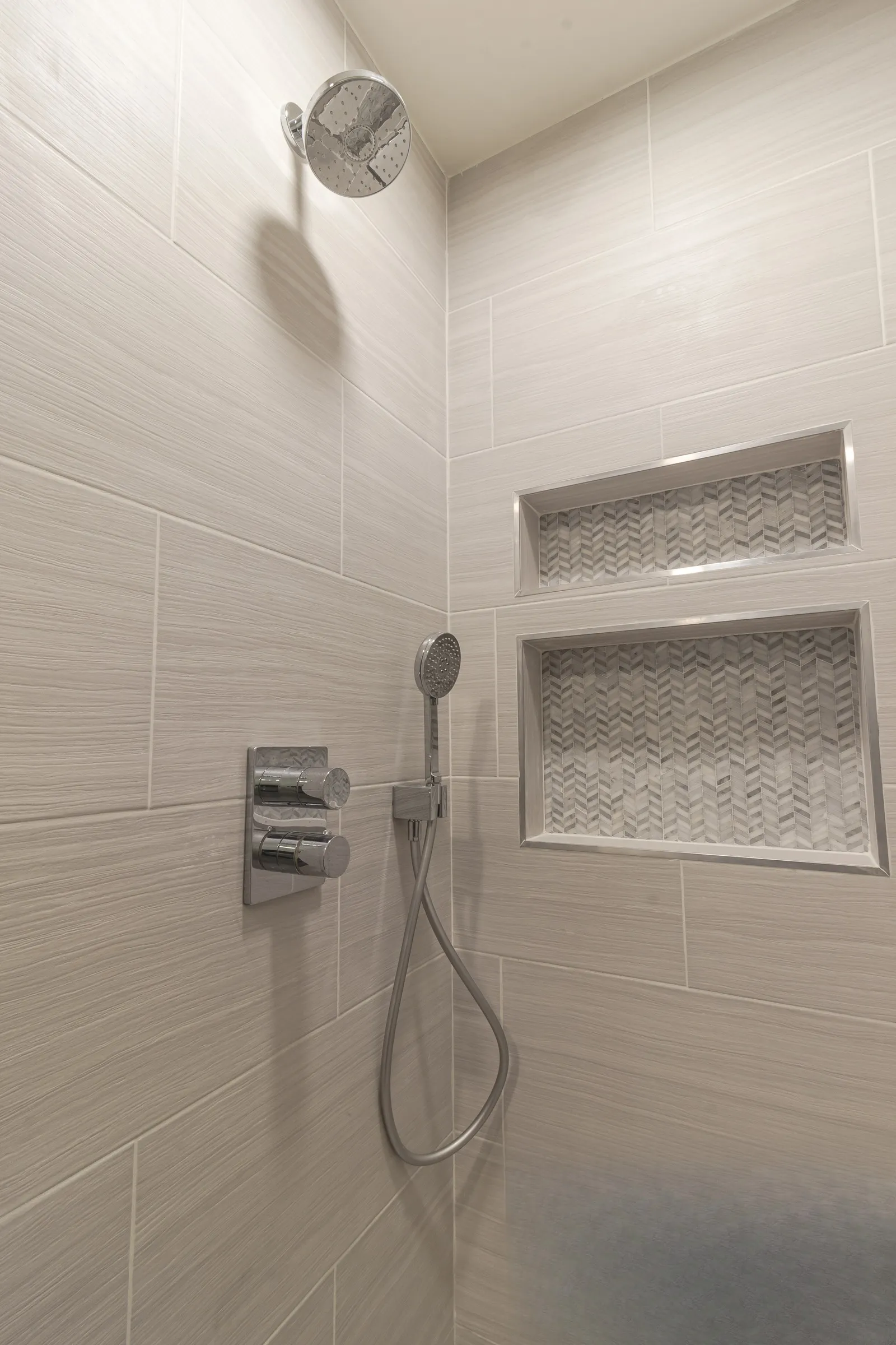 Shower niches lined in herringbone mosaic beside chrome controls and a hand shower