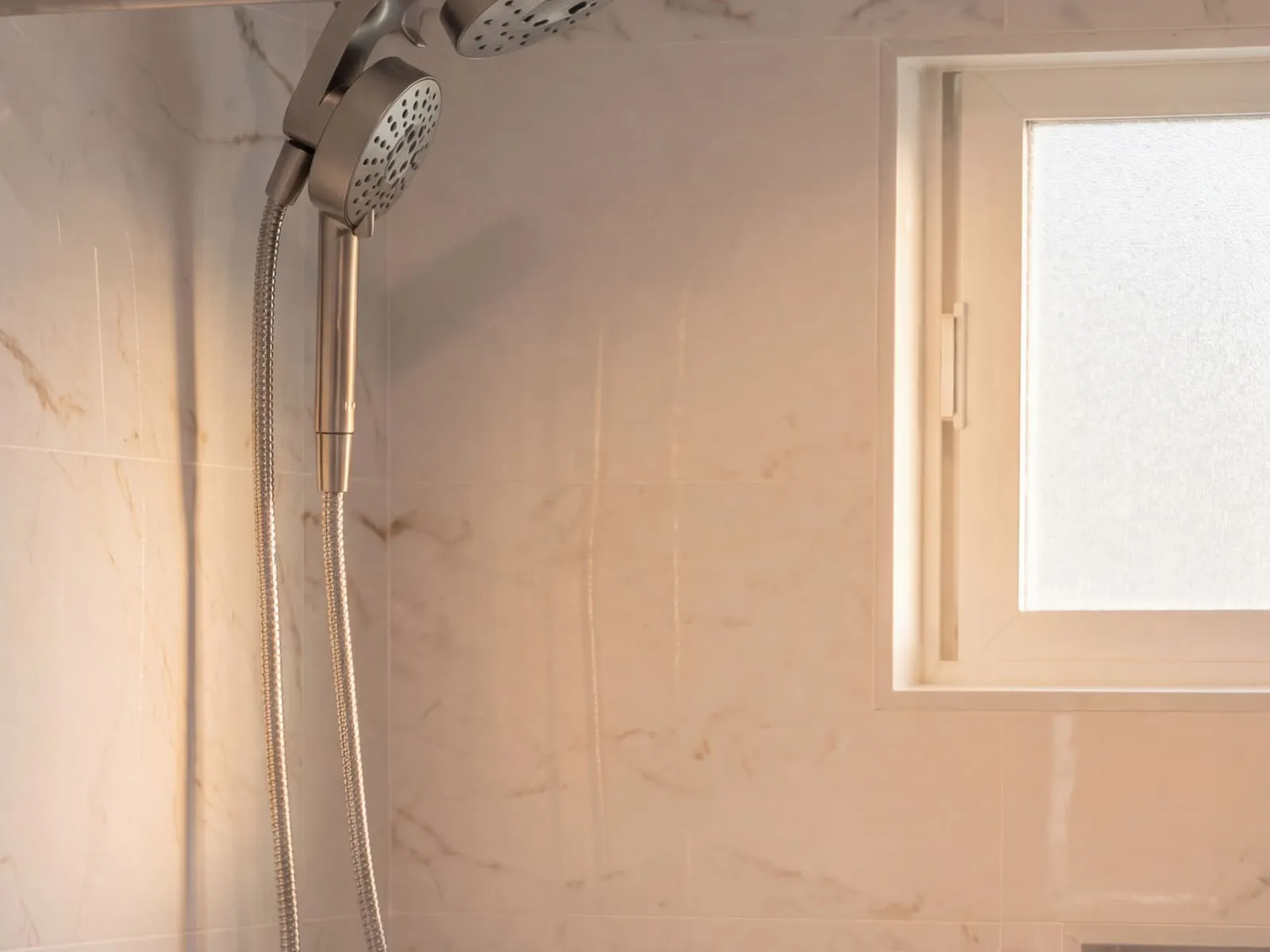 Handheld shower on a slide bar against marble-look tile, beside a frosted window in the tub surround