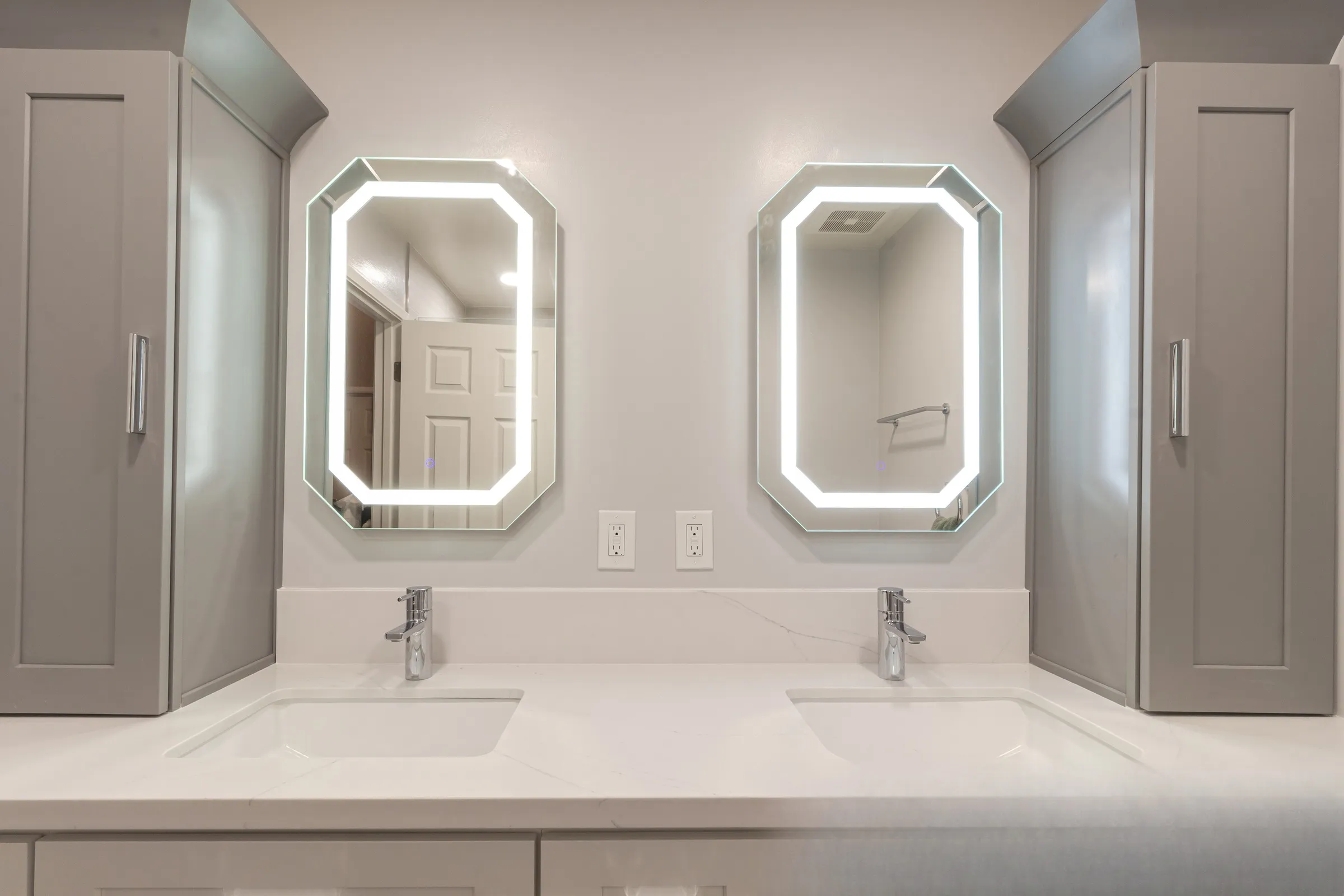 Twin octagonal lit mirrors between two grey shaker linen towers above a veined quartz counter