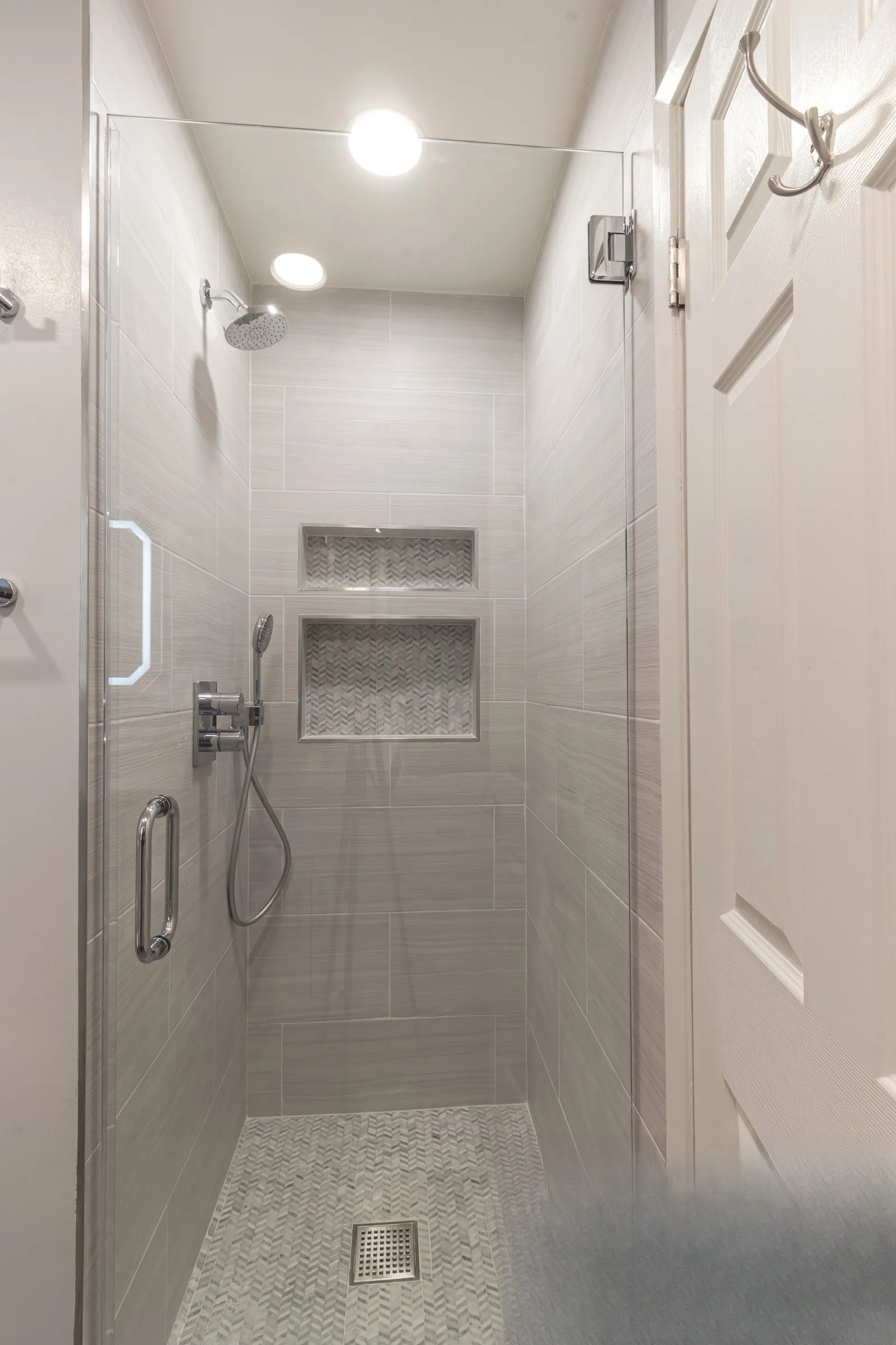 Walk-in shower with glass panel, two recessed niches lined in herringbone mosaic and a herringbone mosaic floor